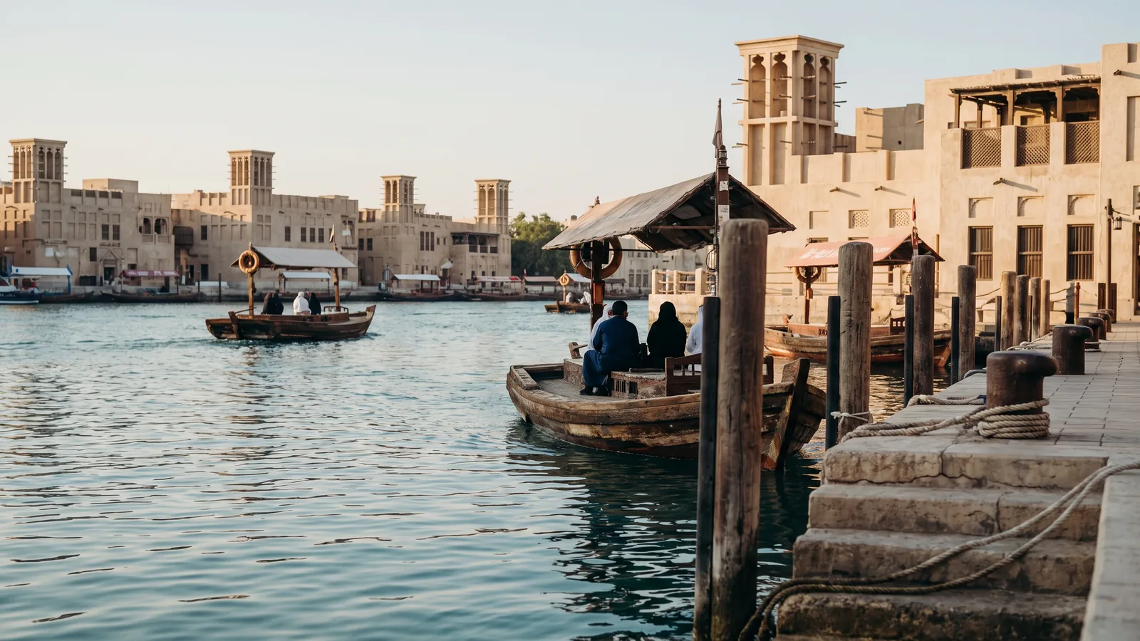 Traditionelle hölzerne Abras auf dem ruhigen Wasser des Dubai Creek vor historischen Sandsteingebäuden mit Windtürmen im Sonnenlicht.