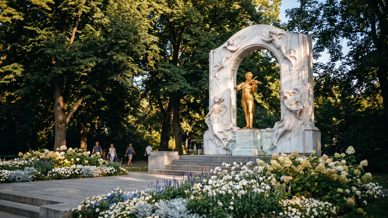 Das vergoldete Johann-Strauß-Denkmal im grünen Wiener Stadtpark umgeben von gepflegten Blumenbeeten im warmen Augustlicht.
