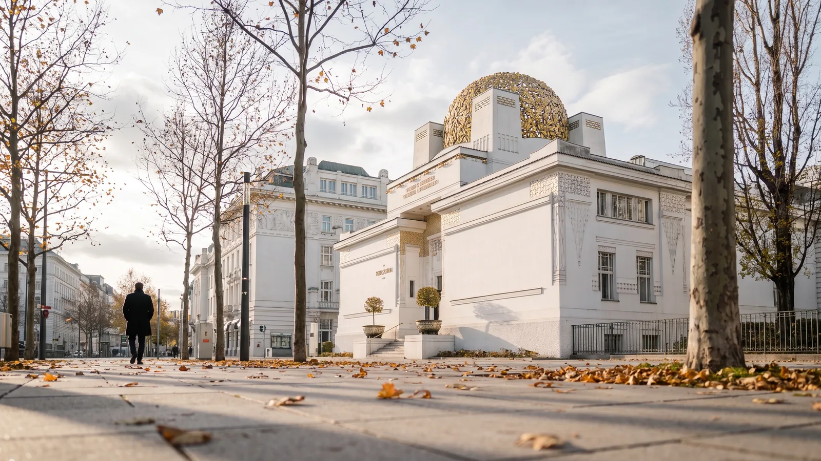 Die weiße Fassade der Wiener Secession mit ihrer vergoldeten Lorbeerkuppel im kühlen Spätherbstlicht neben herbstlichen Bäumen.