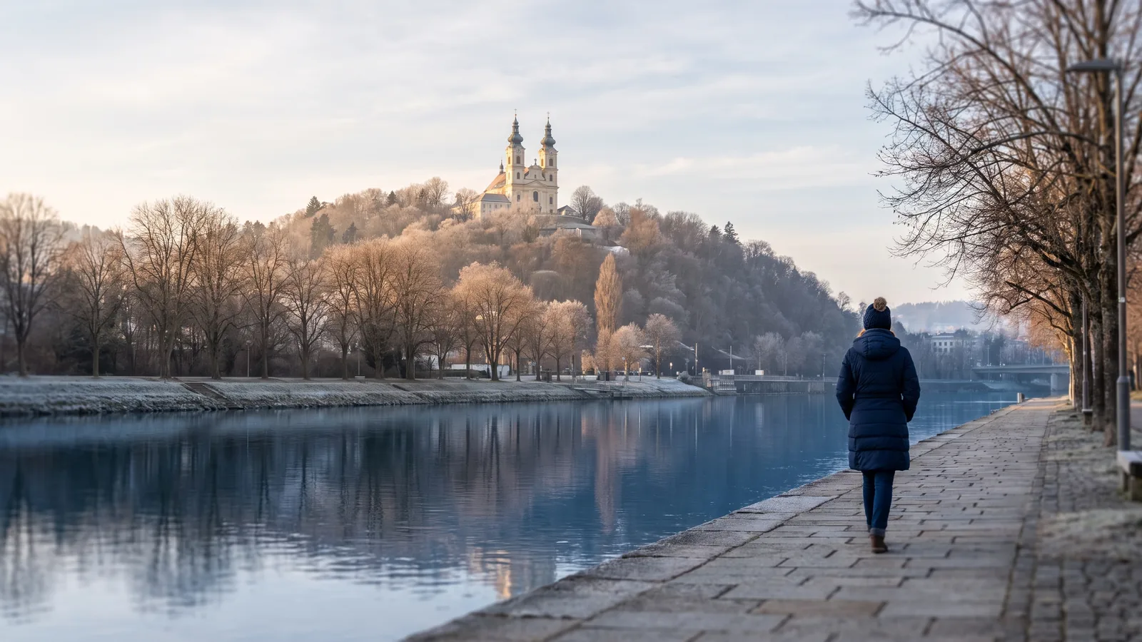 Blick über das ruhige Wasser der Donau auf den bewaldeten Pöstlingberg mit seiner barocken Basilika im Winterlicht.