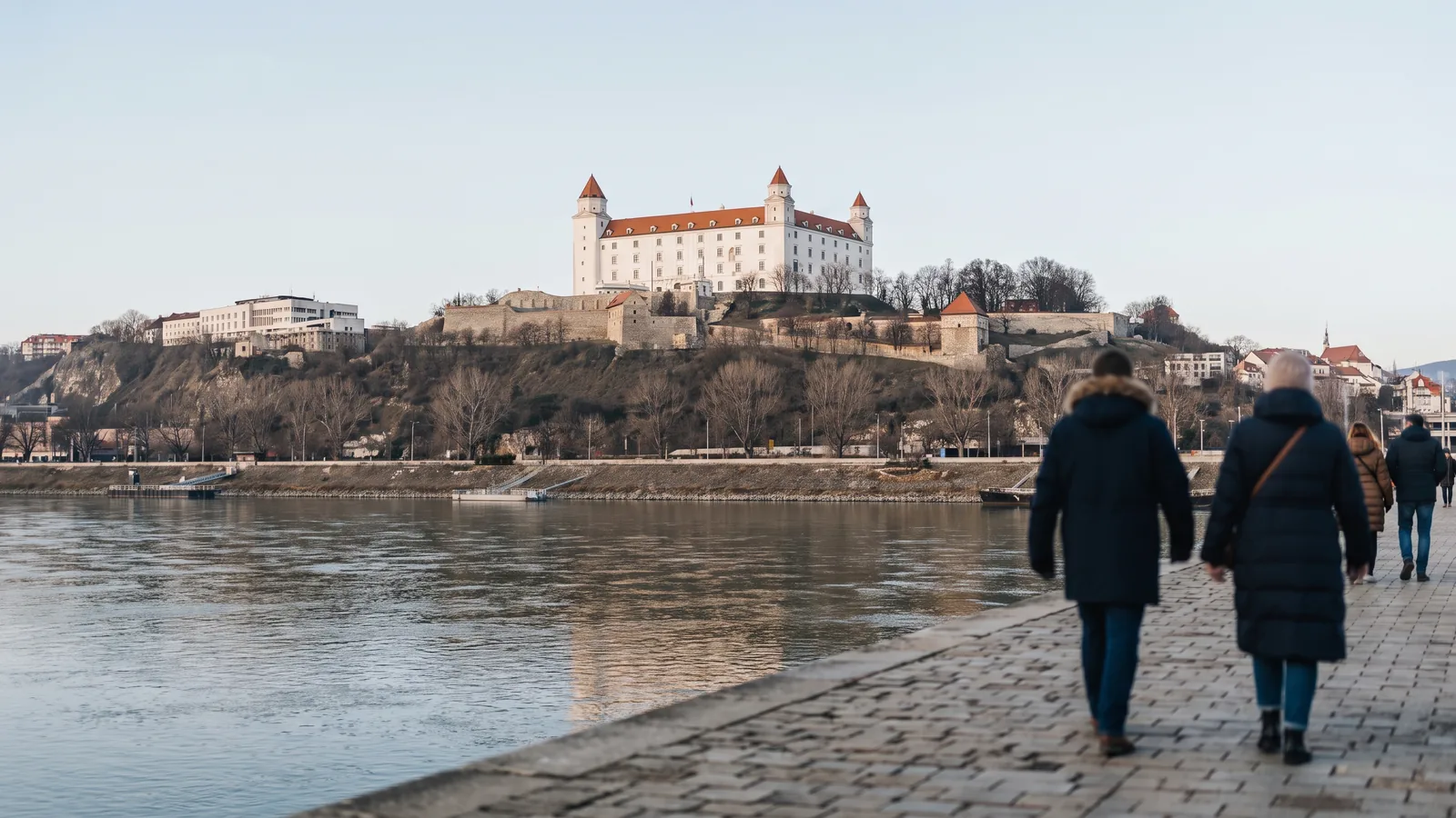Blick über die Donau auf die weiße Burg von Bratislava auf einem Hügel im klaren Winterlicht.